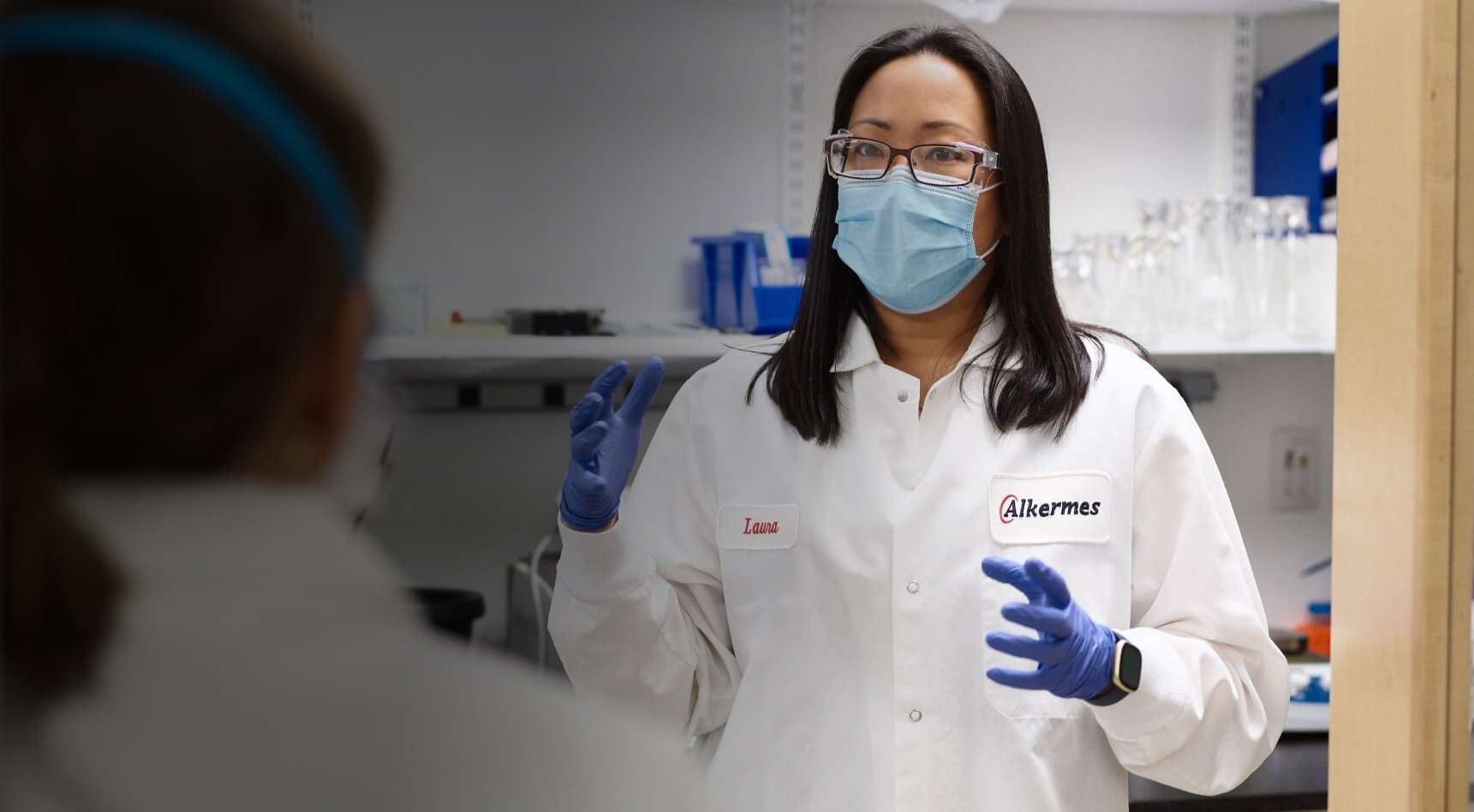 Woman in lab coat with blue gloves, glasses and a mask speaking to colleague at Alkermes